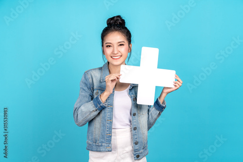Happy asian woman standing and holding plus or add sign on blue background. Cute asia girl smiling wearing casual jeans shirt and showing join sign for increse, upgrade and more benefit concept photo
