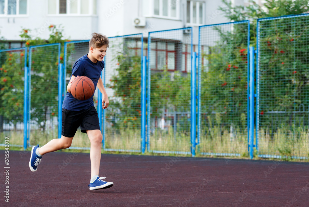 Cute boy in blue t shirt plays basketball on city playground. Active teen enjoying outdoor game with orange ball. Hobby, active lifestyle, sport for kids. 