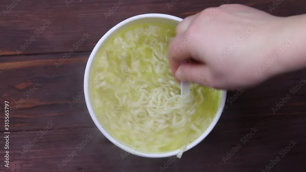 Close-up. A hand with a white plastic fork stirs the noodles. Capacity of paper. Soup is brewing. Close-up. A hand with a white plastic fork stirs the noodles. Capacity of paper. Soup is brewing.