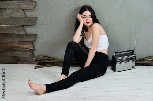 The concept of a glamorous female portrait. Portrait of a pretty brunette girl with long hair with excellent makeup in jeans and a T-shirt on a gray background sitting on the floor near the wall.