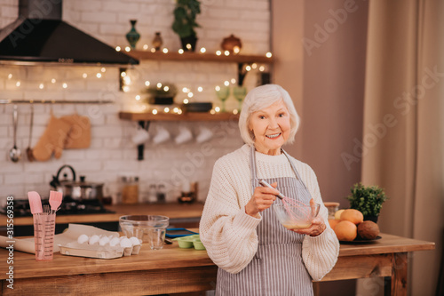 Grey-haired smiling lady standing near the table in the kitchen