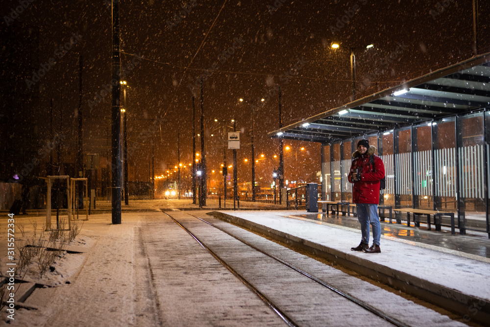 man in red winter coat standing at bus tram stop waiting for public transport
