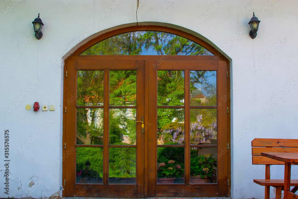 Wooden front door of a rural white house with glass reflecting the green garden and two lamp lights on both sides