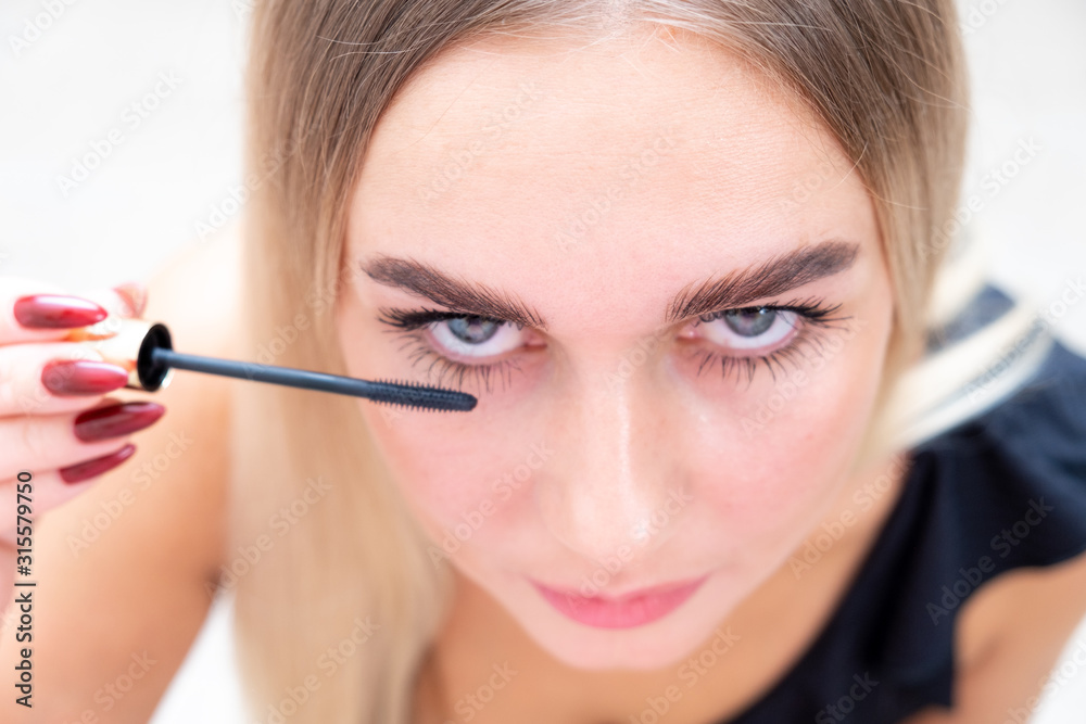 Woman puts black mascara on her eyelashes with makeup, photo of attractive blonde on a white and gray background. Woman puts black mascara on her eyelashes with makeup, photo of attractive blonde on a white and gray background.