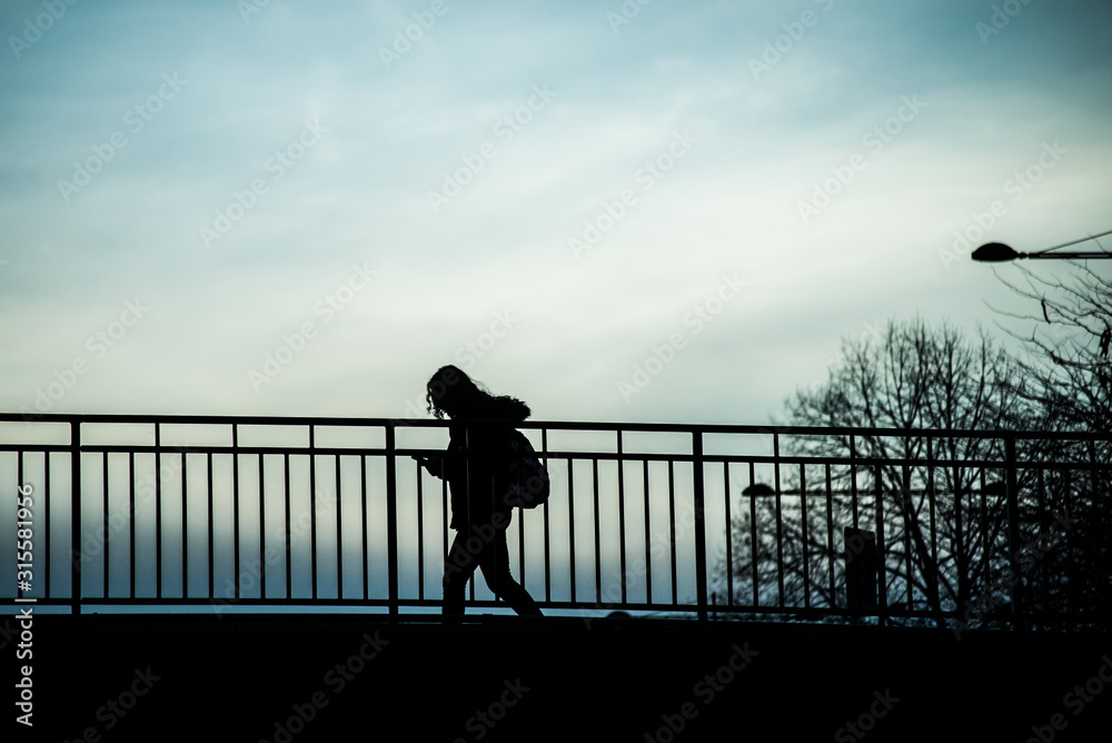 Silhouette of girl walking on bridge under the river with smartphone in hands by sunset