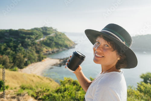 Close-up portrait of a Cute Woman in a Hat with a paper cup of coffee in hand on the background of a luxurious landscape of the sea, mountains and the beach. Thailand.