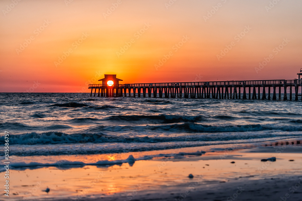 Naples, Florida colorful red sunset in gulf of Mexico with sun path reflection and Pier wooden jetty silhouette with horizon and ocean waves