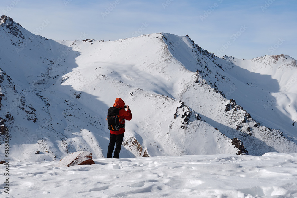 Man in red jacket with black backpack is standing at mountain plateau against the high snowy mountains.