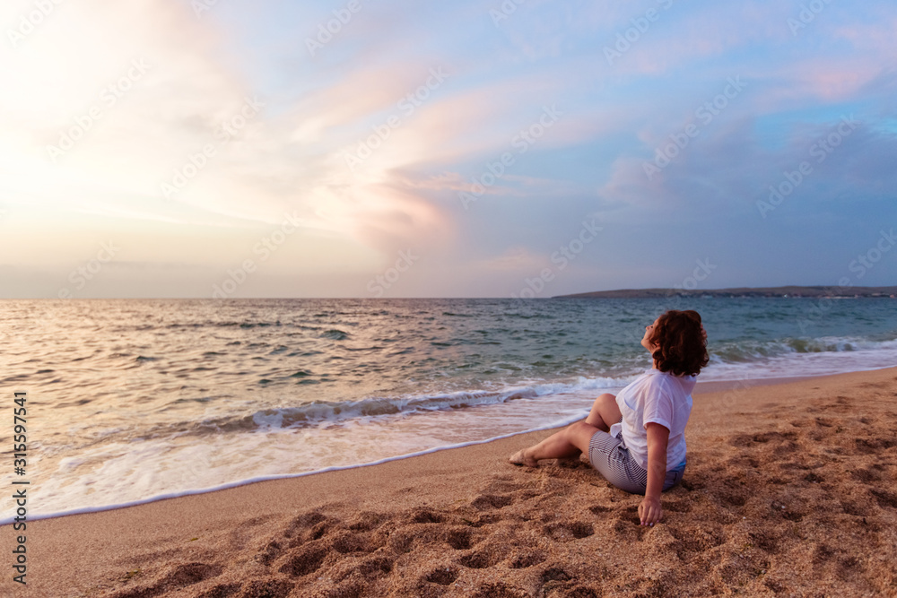 A woman in a white shirt with dark hair sits on the yellow sand of shells on the seashore and looks at the sunset.
