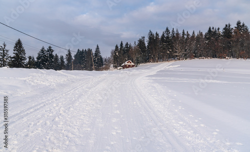 winter scenery with isolated wooden house, snow covered road and meadows, forest and blue sky with clouds photo