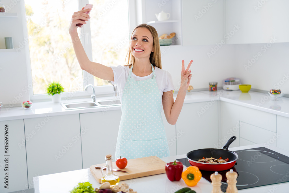 Photo of housewife weekend morning cooking holding telephone showing v-sign presenting followers traditional family recipe frying pan meal white kitchen indoors