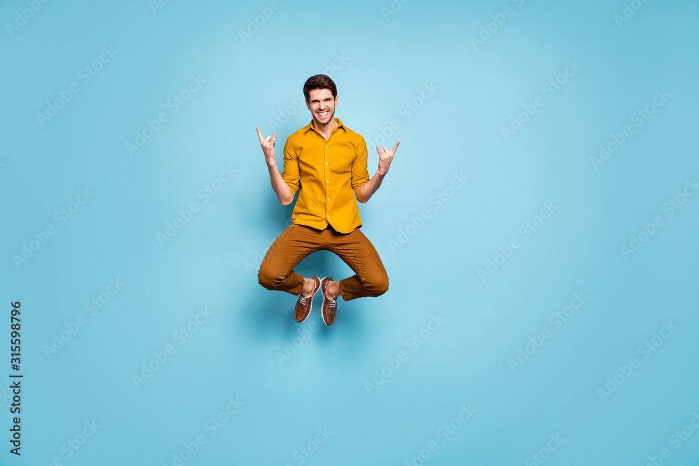 Obraz premium Full length photo of funny guy jumping high raising hands showing horns rejoicing rock concert wear yellow shirt pants isolated blue color background