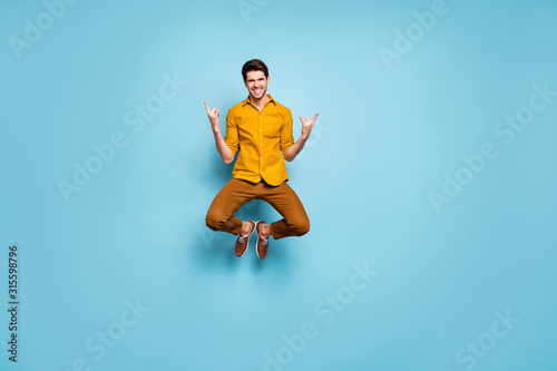 Full length photo of funny guy jumping high raising hands showing horns rejoicing rock concert wear yellow shirt pants isolated blue color background