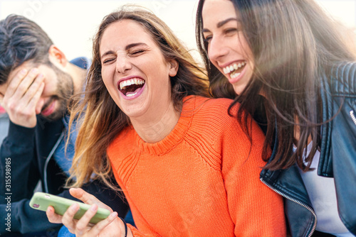 Fotografía Euphoric group of friends laughing with open mouth and joking holding a smartphone in the hand sitting on an outdoors bench