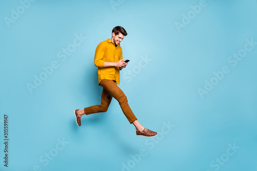 Full length profile photo of funny guy jumping high holding telephone millennial writing email walking down street wear yellow shirt pants isolated blue color background