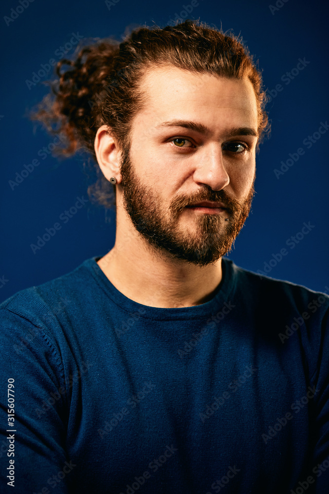 Portrait of gorgeous caucasian bearded hipster in blue shirt standing in front of blue background and having serious facial expression.