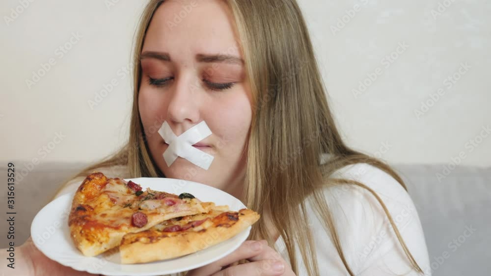 Young woman with white duct tape over her mouth, preventing her to eat junk food. The girl is offered to smell a delicious piece of pizza on a plate. Young woman with white duct tape over her mouth, preventing her to eat junk food. The girl is offered to smell a delicious piece of pizza on a plate.