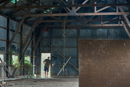 A rope-swing made from a length of building timber, hangs motionless from the wooden trussed ceiling, above the dirty concrete floor of an abandoned industrial warehouse. A man stands looking out.
