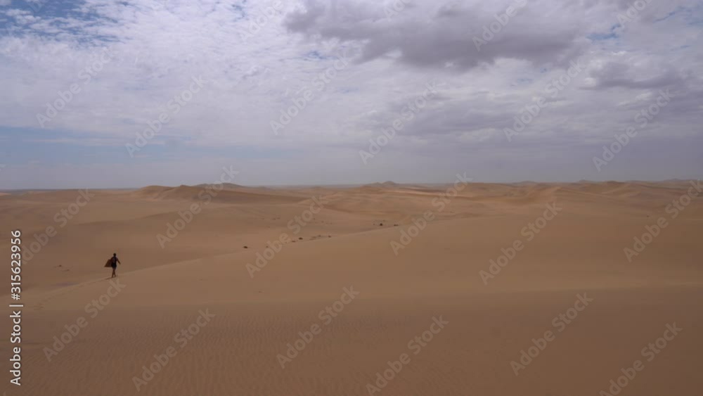 Sandboarder walking on dunes in the Namib-Naukluft National Park on a cloudy day, Namibia. Still wide shot. Sandboarder walking on dunes in the Namib-Naukluft National Park on a cloudy day, Namibia. Still wide shot.