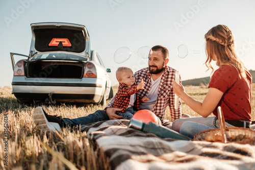 Happy Young Family Mom and Dad with Their Little Son Enjoying Summer Weekend Picnic on the Car Outside the City, Playing with Bubbles