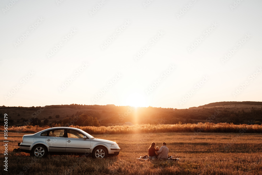Happy Young Family Mom and Dad with Their Little Son Enjoying Summer Weekend Picnic on the Car Outside the City in the Field at Sunny Day Sunset, Vacation and Road Trip Concept