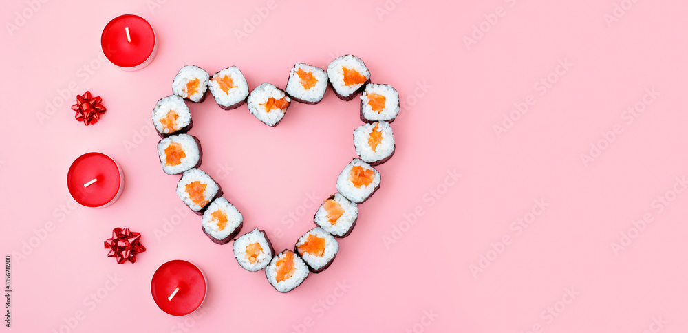 Obraz premium Salmon sushi rolls laid out in the shape of a heart, next to red candles and bowson a pink background. The concept of Japanese cuisine for Valentine's Day, greeting card, banner. Copy space
