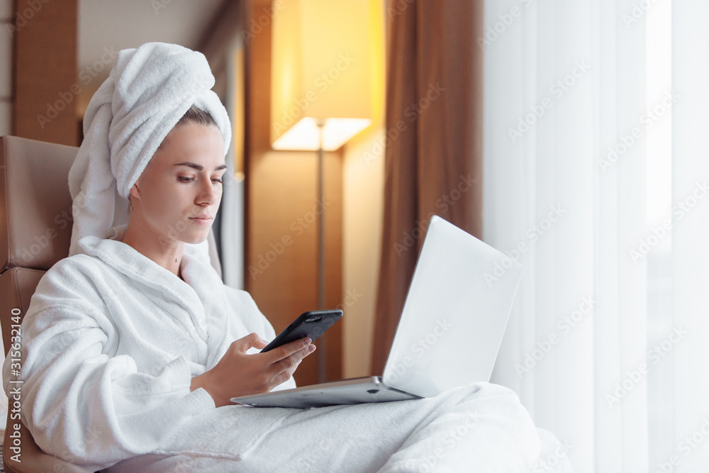 Attractive young business woman chatting on social networks using smartphone with boyfriend resting after a shower in a towel and bathrobe in a hotel room working with a laptop. Working travel concept