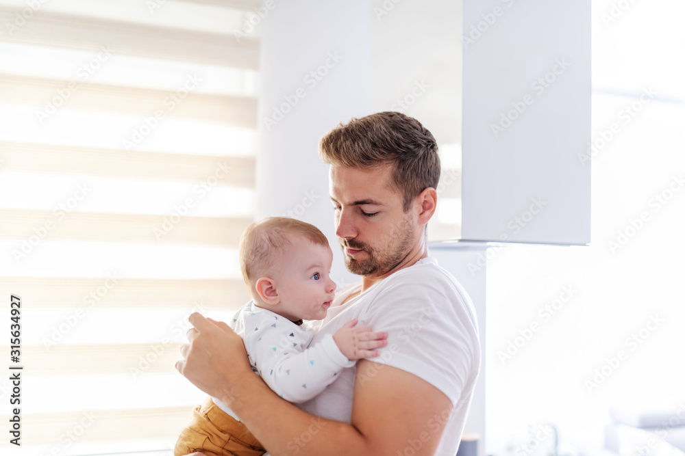 Side view of caring handsome caucasian young dad holding his loving 6 months old baby boy while standing next to window. Unconditional love concept. Side view of caring handsome caucasian young dad holding his loving 6 months old baby boy while standing next to window. Unconditional love concept.