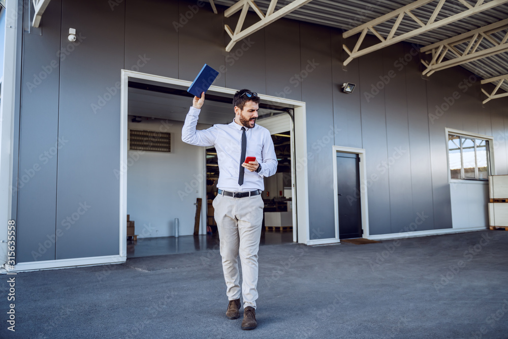 Angry Caucasian director walking in front of printing shop, holding smart phone and notebook and swearing. Angry Caucasian director walking in front of printing shop, holding smart phone and notebook and swearing.