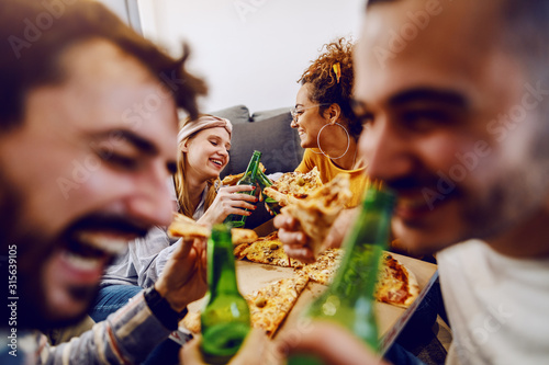 Group of friends sitting on the floor in living room, drinking beer and eating pizza. House party.