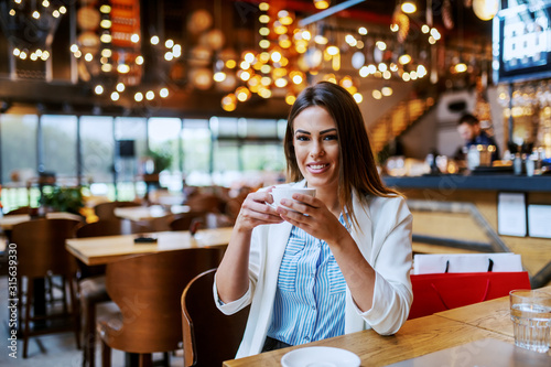 Gorgeous fashionable caucasian brunette with beautiful smile sitting in coffee shop and holding cup of fresh delicious espresso. Behind her backs on chair are shopping bags.