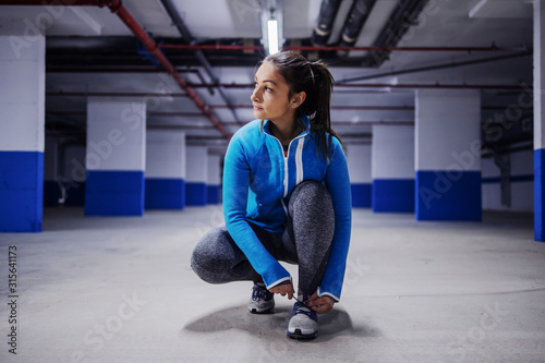 Young fit attractive caucasian sportswoman in tracksuit crouching and tying shoelace. Urban life concept.