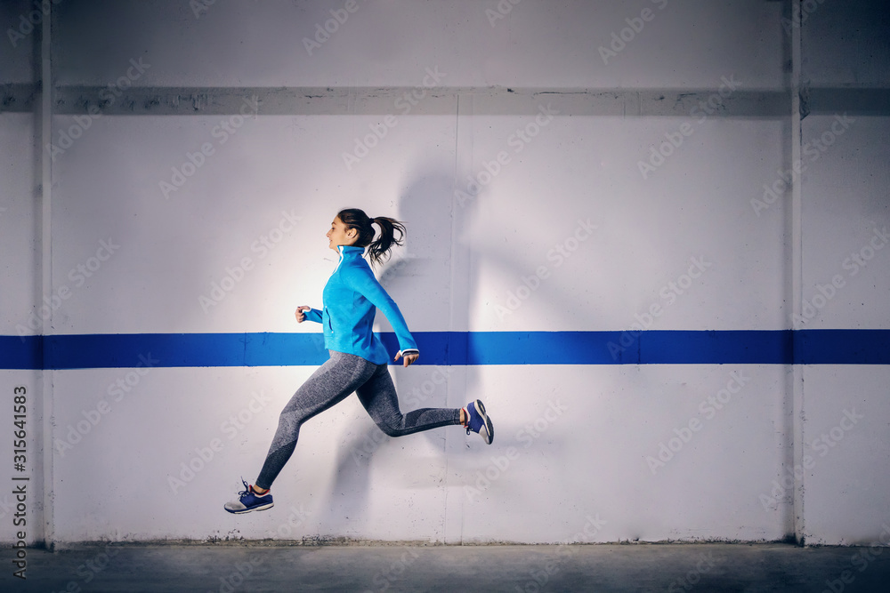 Young fit attractive caucasian sportswoman in tracksuit running fast in garage. Urban life concept.