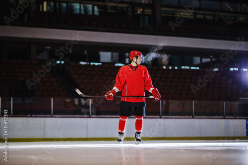 Hockey player in uniform and with stick in hands standing on ice and preparing to attack opposite player. Hall interior.