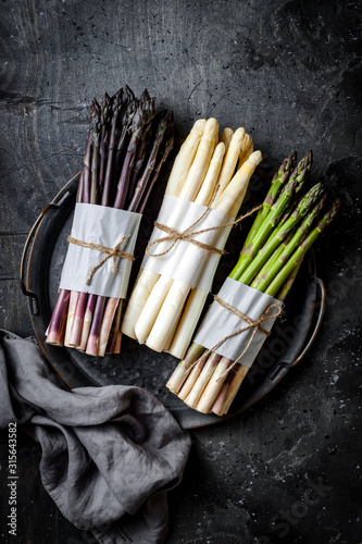 Bunches of fresh green, purple, white asparagus on vintage metal tray over dark grey rustic background. Top view, copy space
