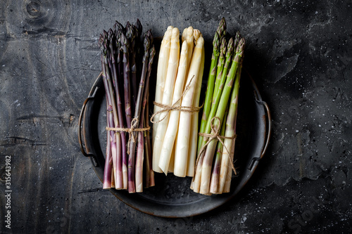 Bunches of fresh green, purple, white asparagus on vintage metal tray over dark grey rustic background. Top view, copy space