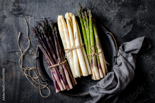 Bunches of fresh green, purple, white asparagus on vintage metal tray over dark grey rustic background. Top view, copy space photo