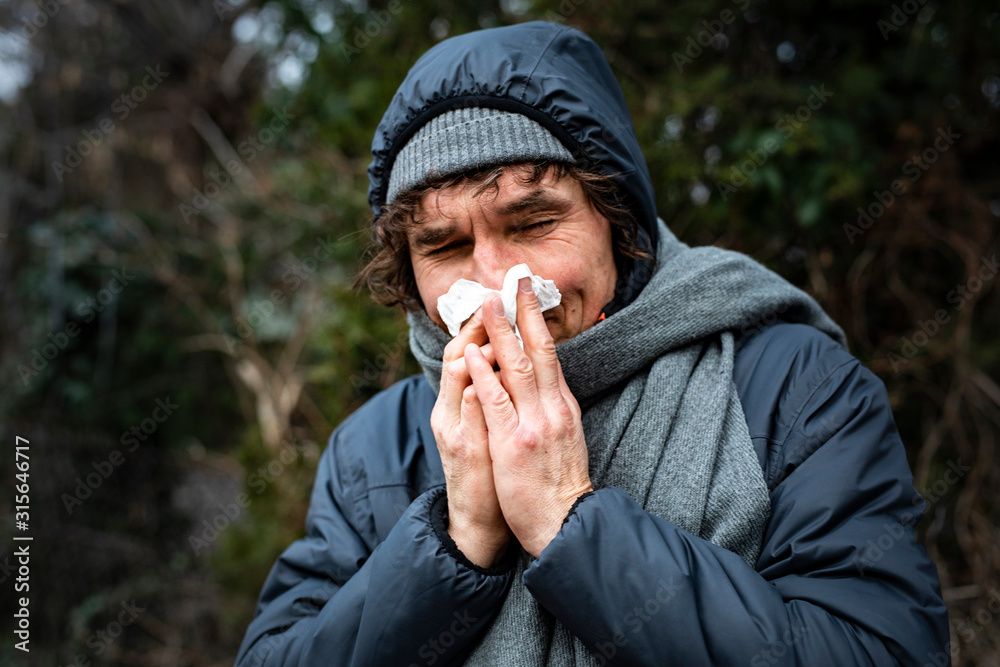 Man blowing his nose with a tissue when walking outdoor. Runny nose, infection, flu, viral diseases. - obrazy, fototapety, plakaty 