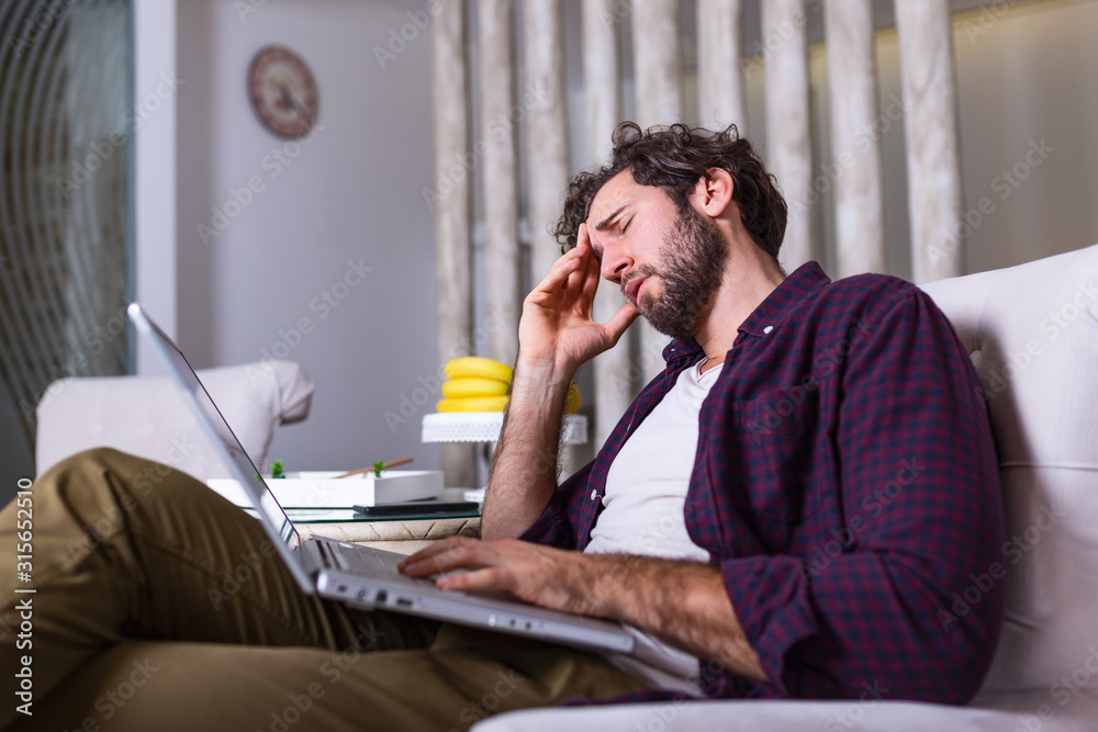 Young tired manager sitting at home surrounded with papers and laptop computer having tired look holding his hand on head having pain closing his eyes being sleepy and exhausted. Tiredness Young tired manager sitting at home surrounded with papers and laptop computer having tired look holding his hand on head having pain closing his eyes being sleepy and exhausted. Tiredness