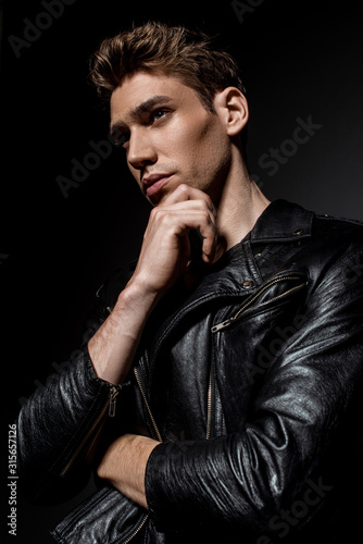 sexy young man in biker jacket touching chin and looking away on black background