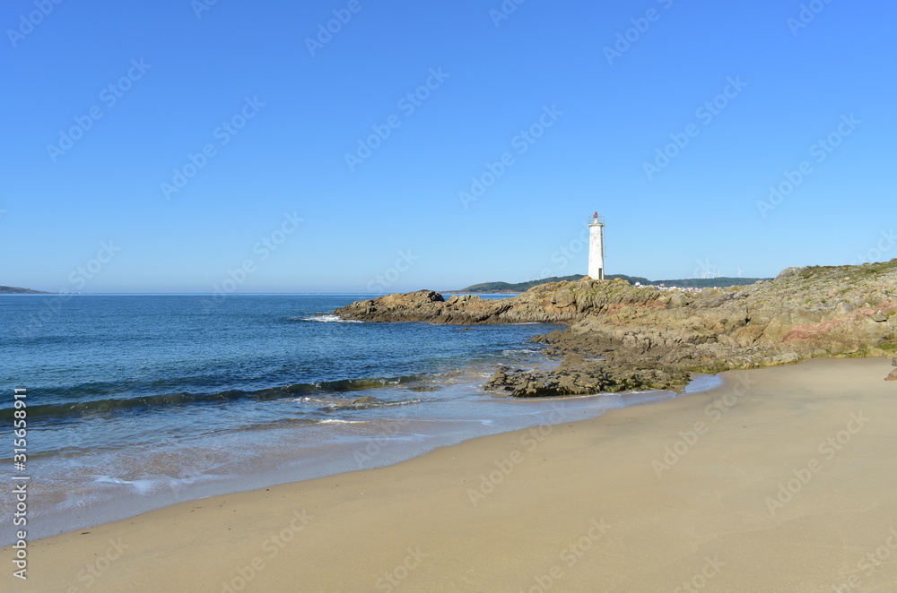 Beach with wet sand, rocks and lighthouse with blue sky. Muxia, Coruña, Galicia, Spain.