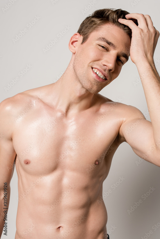 smiling sexy man with muscular torso touching hair isolated on grey