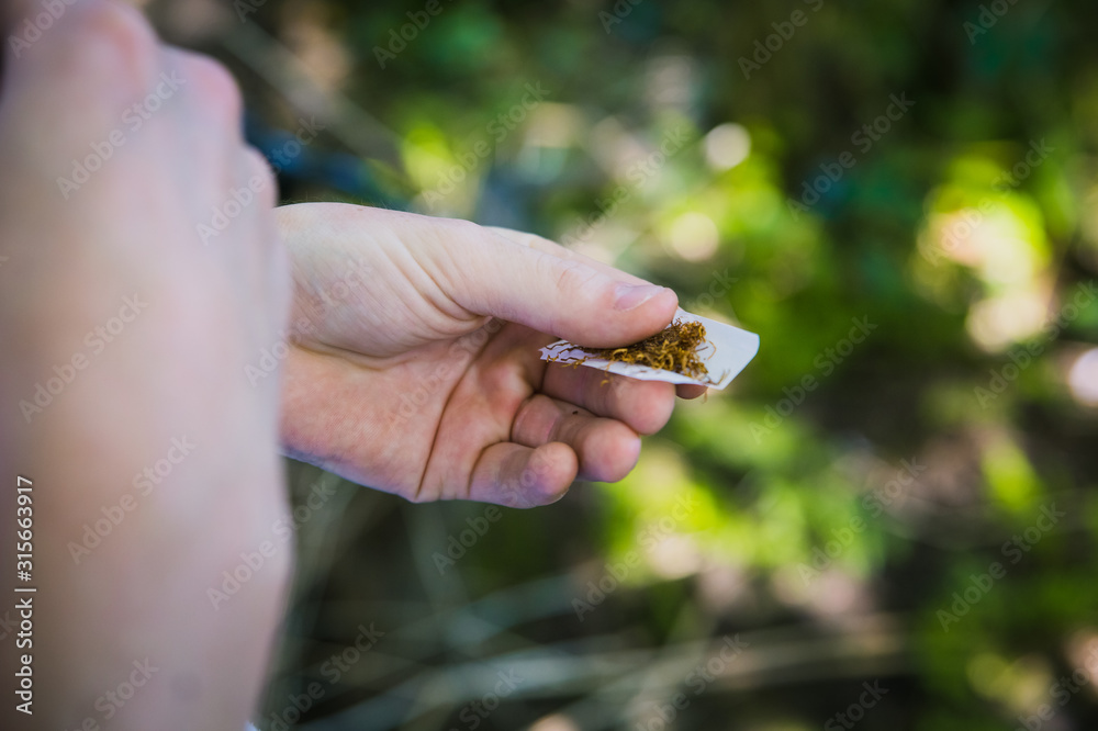 Man under the influence of drugs or alcohol struggling to roll a tobacco cigarette or marijuana joint or spliff in the wood forest with nice sunny green bokeh background pale young hands
