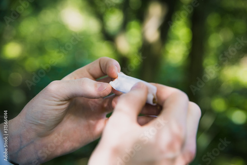 Man under the influence of drugs or alcohol struggling to roll a tobacco cigarette or marijuana joint or spliff in the wood forest with nice sunny green bokeh background pale young hands © josh