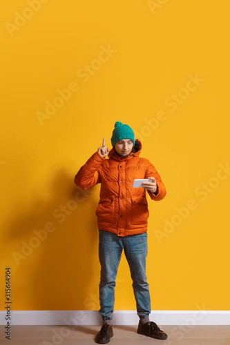 Pointing up. Young caucasian man using smartphone, serfing, chatting, betting. Full length portrait isolated on yellow background. Concept of modern technologies, millennials, social media.