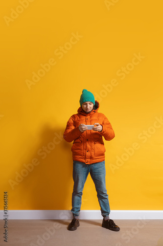 Playing with gadget. Young caucasian man using smartphone, serfing, chatting, betting. Full length portrait isolated on yellow background. Concept of modern technologies, millennials, social media.