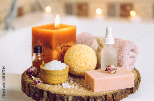 Fotomurale Home spa products on wooden disc tray: bar of soap, bath bomb, aroma bath salt, essential and massage oils, candle burning, rolled towel inside bathroom by tub, water running