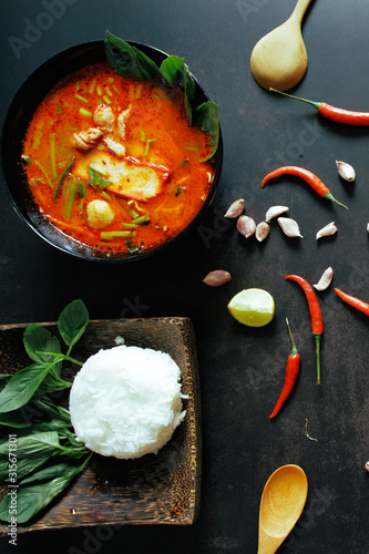 Thai food vertical background concept. Dish of Thailand cuisine. Tom yum pork soup in black dish, rice in wooden plate, wooden ladle and spoon, chilis, garlic on table dark background