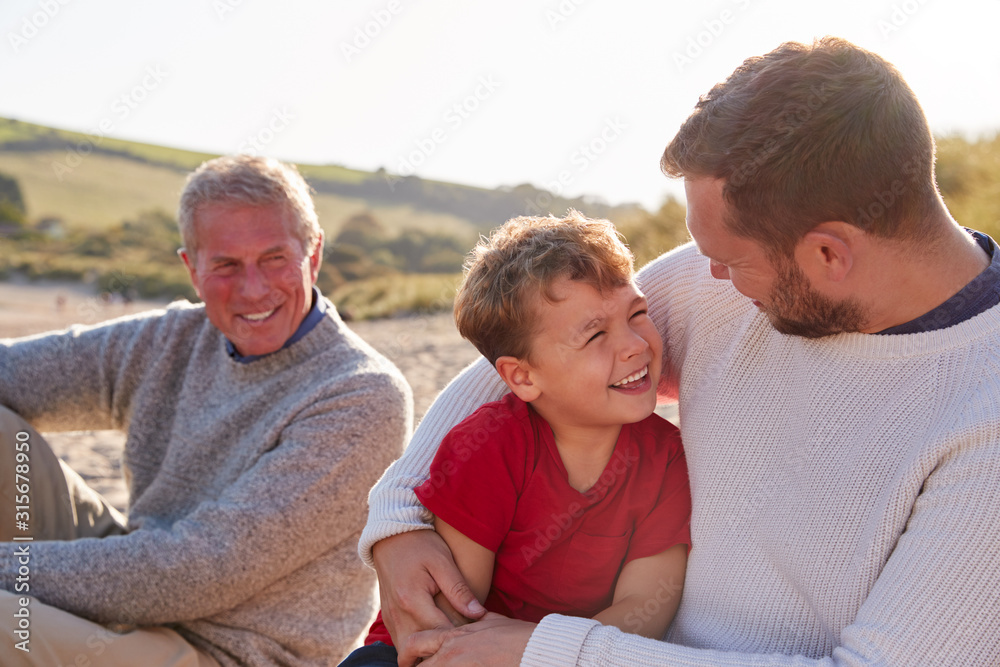 Family With Grandfather Relaxing On Beach With Adult Son And Grandson