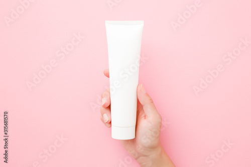 Young woman hand holding and showing white tube on pastel pink table. Care about clean and soft body skin. Daily beauty product. Closeup. Top down view. Point of view shot.